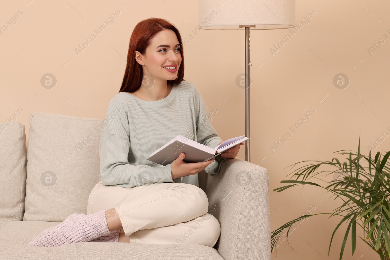 Photo of Happy woman with red dyed hair and book sitting on sofa indoors