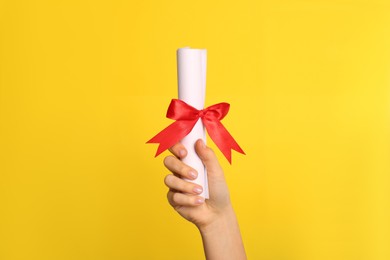 Photo of Student holding rolled diploma with red ribbon on yellow background, closeup