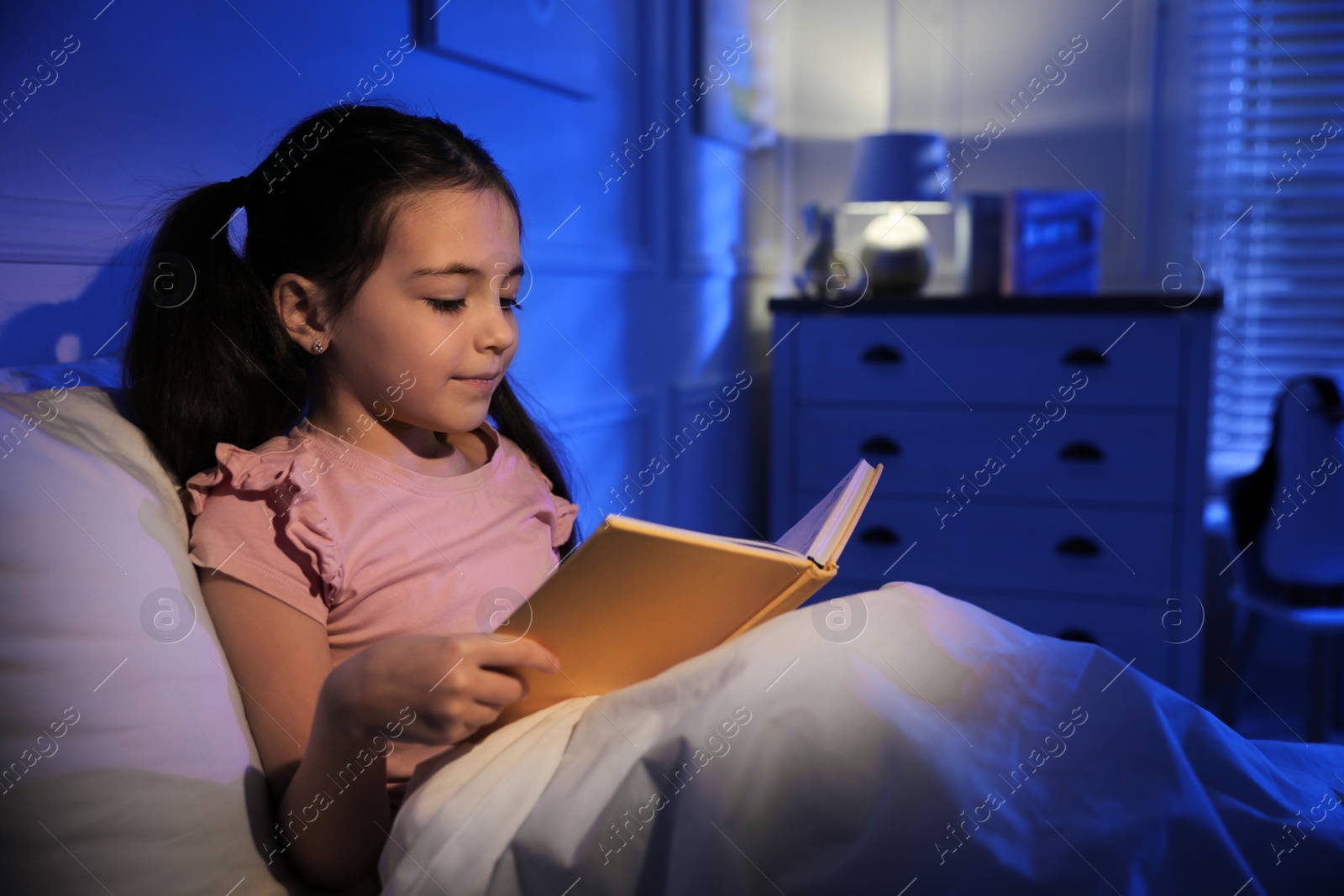 Photo of Little girl reading fairy tale in dark bedroom