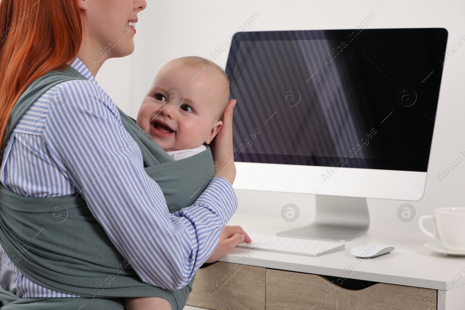 Photo of Mother holding her child in sling (baby carrier) while using computer at workplace