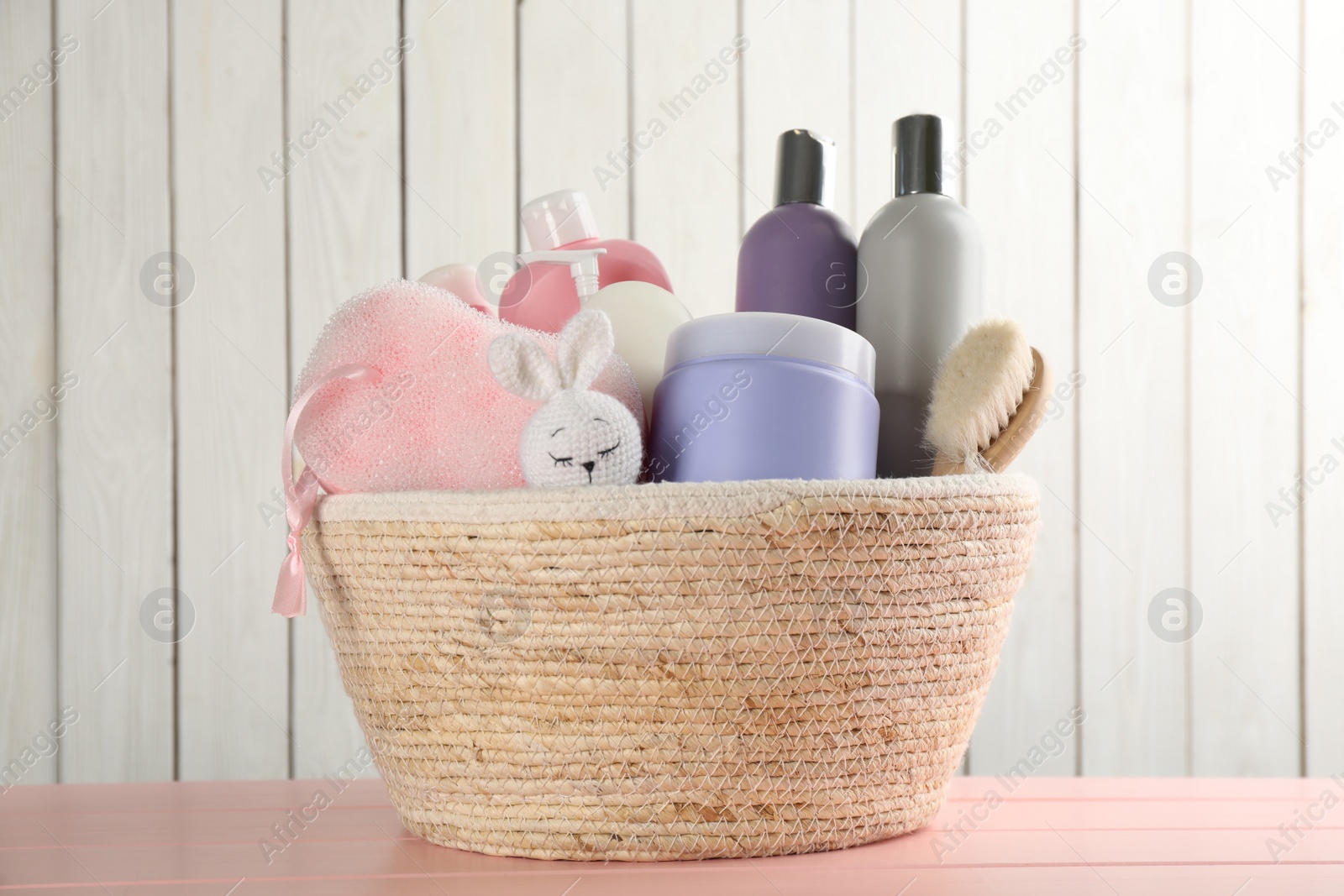 Photo of Wicker basket full of different baby cosmetic products, accessories and toy on pink wooden table