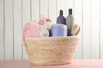 Photo of Wicker basket full of different baby cosmetic products, accessories and toy on pink wooden table