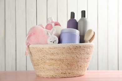 Wicker basket full of different baby cosmetic products, accessories and toy on pink wooden table