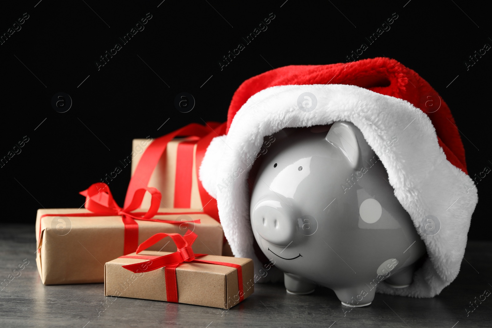 Photo of Piggy bank with Santa hat and gift boxes on grey table against black background