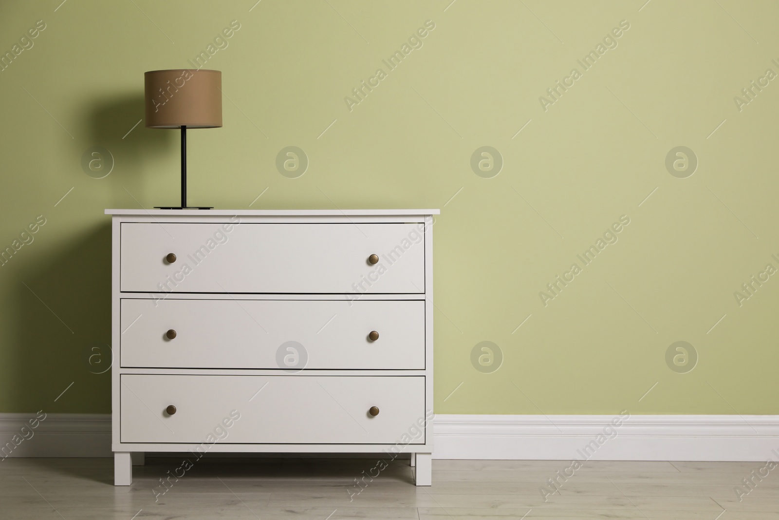 Photo of Modern white chest of drawers with lamp near light green wall indoors. Space for text