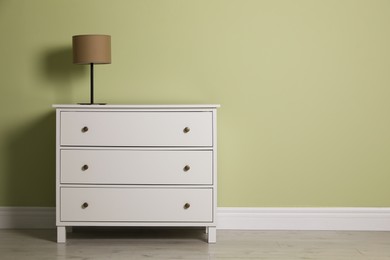 Photo of Modern white chest of drawers with lamp near light green wall indoors. Space for text