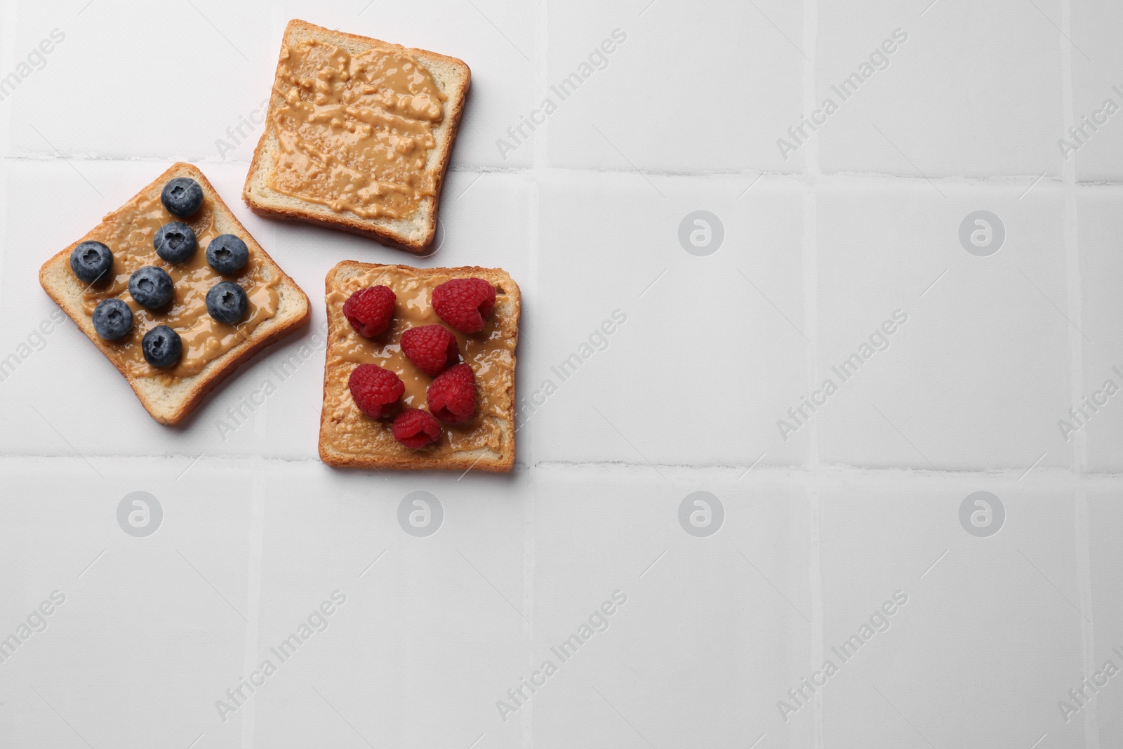 Photo of Delicious toasts with peanut butter, raspberries and blueberries on white tiled table, flat lay. Space for text