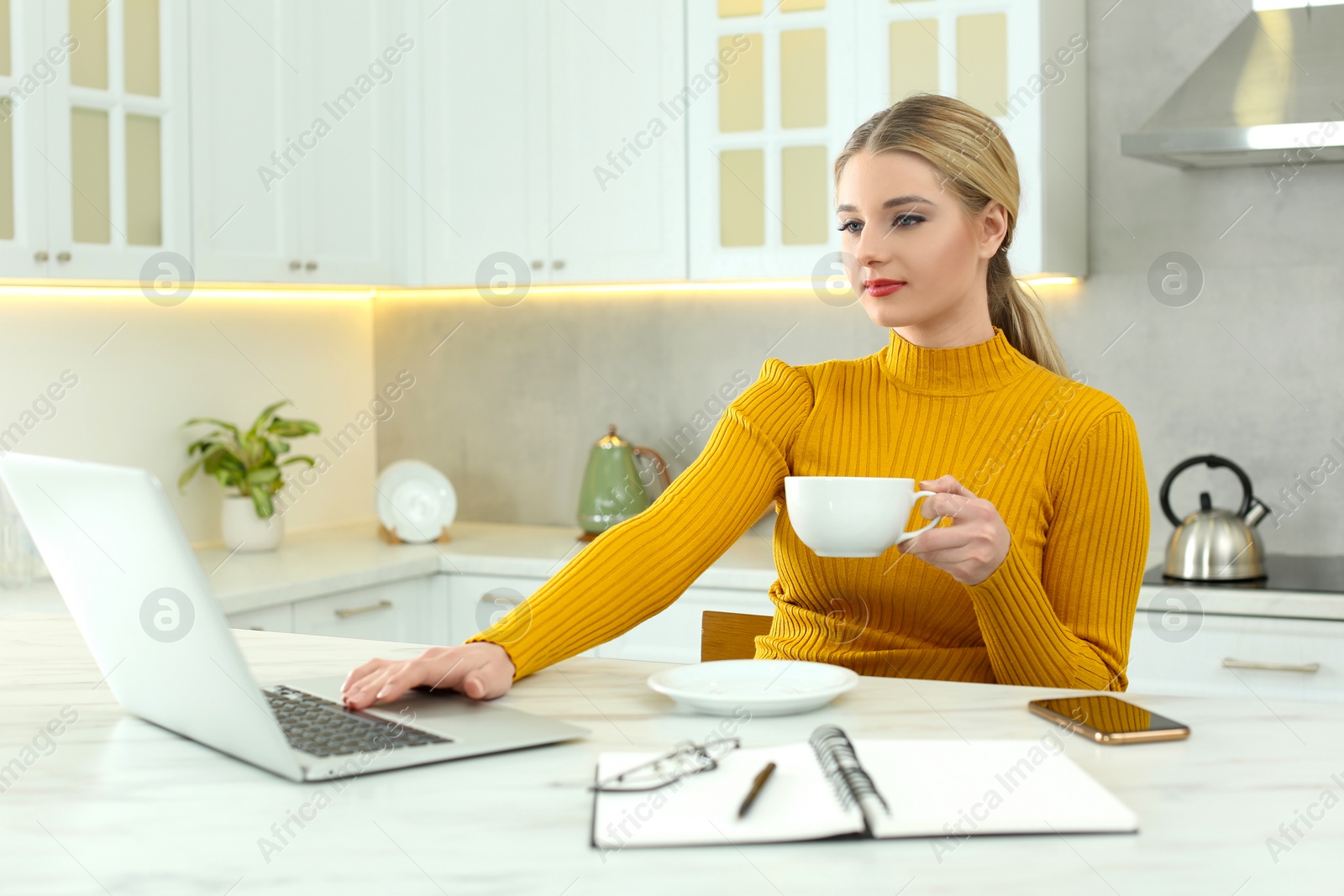 Photo of Home workplace. Woman with cup of hot drink looking at laptop at marble desk in kitchen