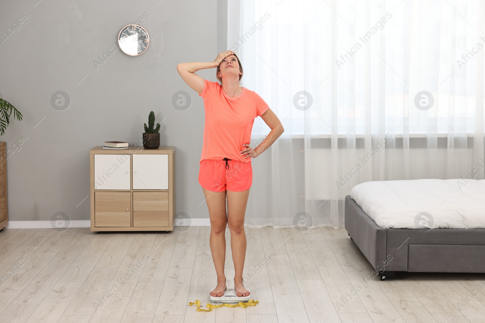 Photo of Unhappy woman in sportswear standing on floor scales at home