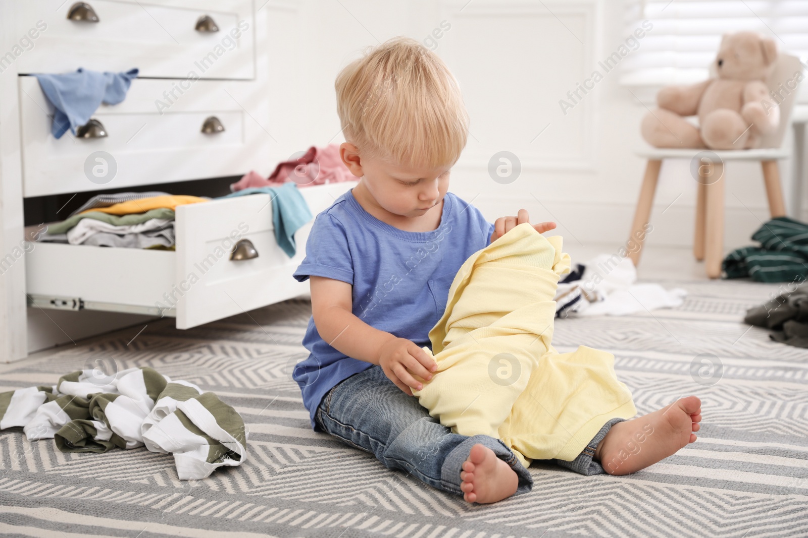 Photo of Cute little boy playing with clothes near dresser on floor at home