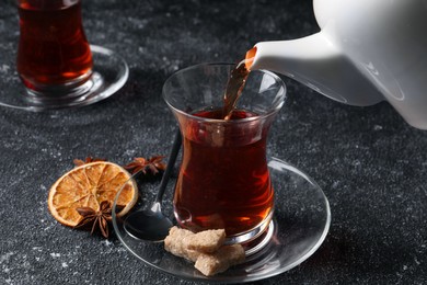 Pouring traditional Turkish tea from pot into glass on grey textured table, closeup