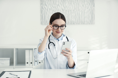 Young female doctor with smartphone at table in office