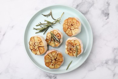 Photo of Heads of fried garlic and rosemary on white marble table, top view