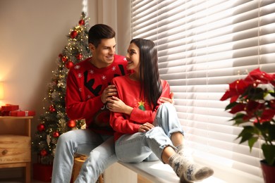Photo of Happy couple in warm Christmas sweaters near window at home
