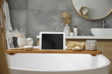 Photo of Wooden tray with tablet and spa products on bath tub in bathroom