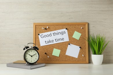 Photo of Cork board with motivational quote Good Things Take Time, notebook, alarm clock and plant on white wooden table