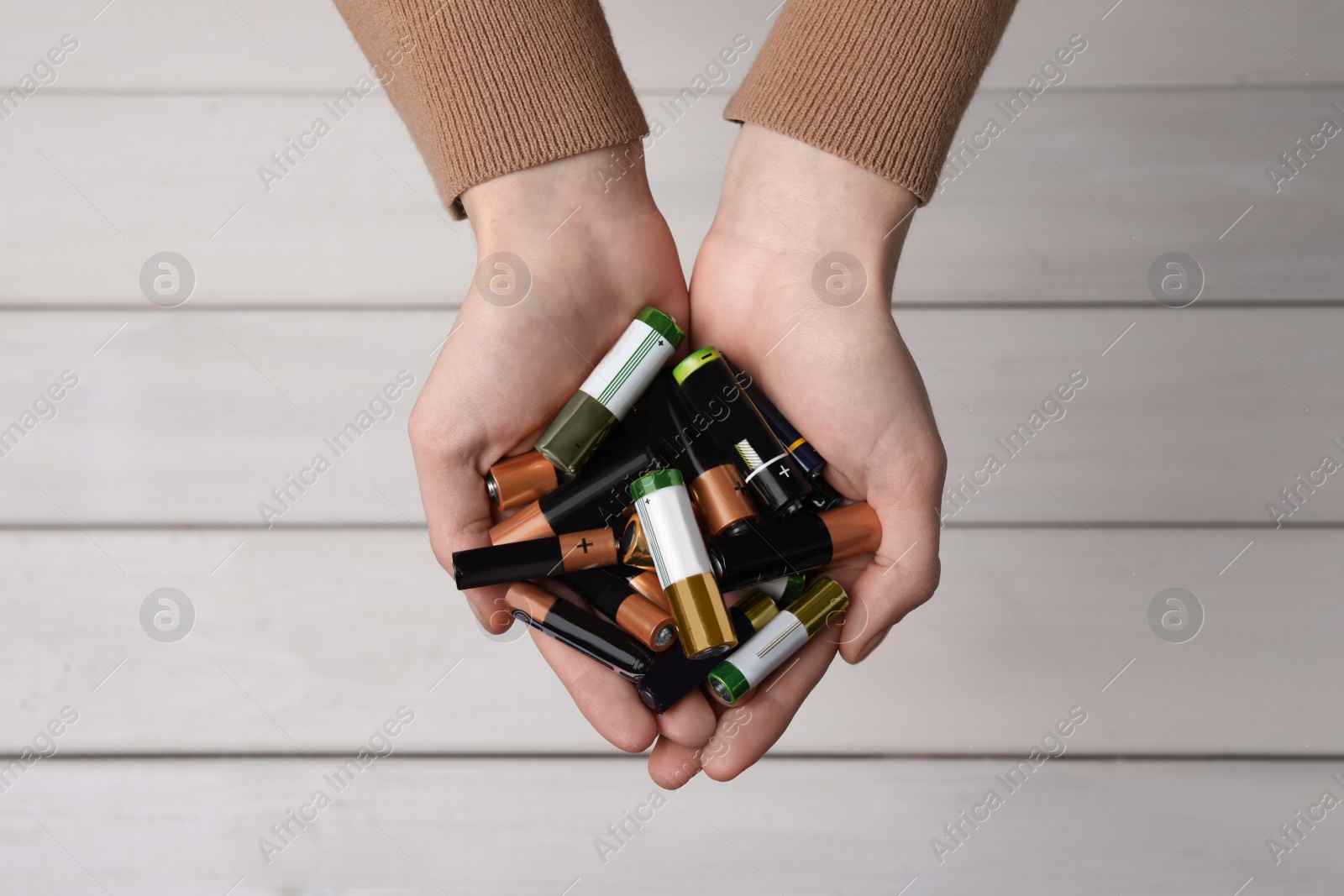Image of Woman holding many used electric batteries in her hands over white wooden table, top view