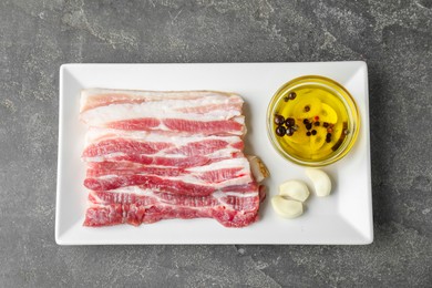 Pieces of raw pork belly, oil and garlic on grey textured table, top view