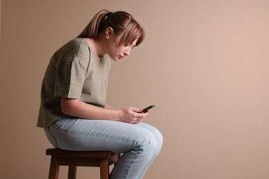 Photo of Young woman with poor posture using smartphone while sitting on stool against beige background, space for text