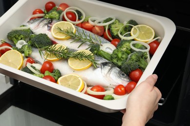 Photo of Woman putting baking dish with raw fish and vegetables into oven, closeup