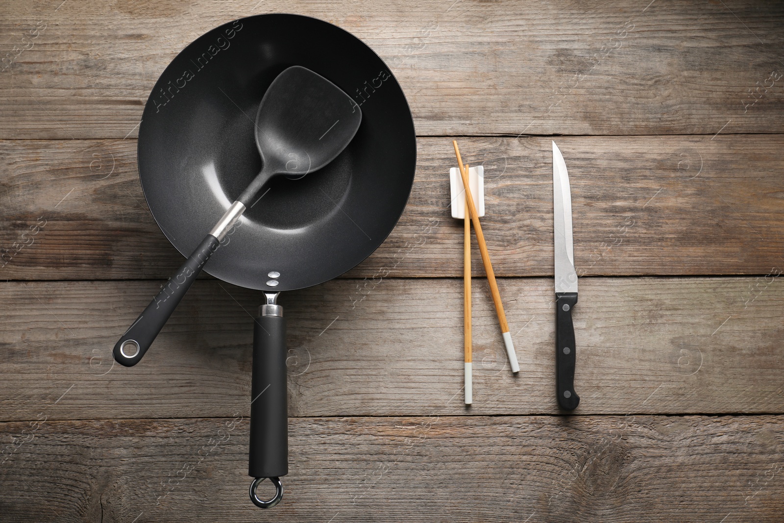 Photo of Black metal wok, chopsticks, knife and spatula on wooden table, flat lay