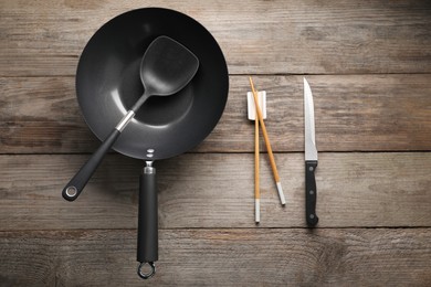 Photo of Black metal wok, chopsticks, knife and spatula on wooden table, flat lay