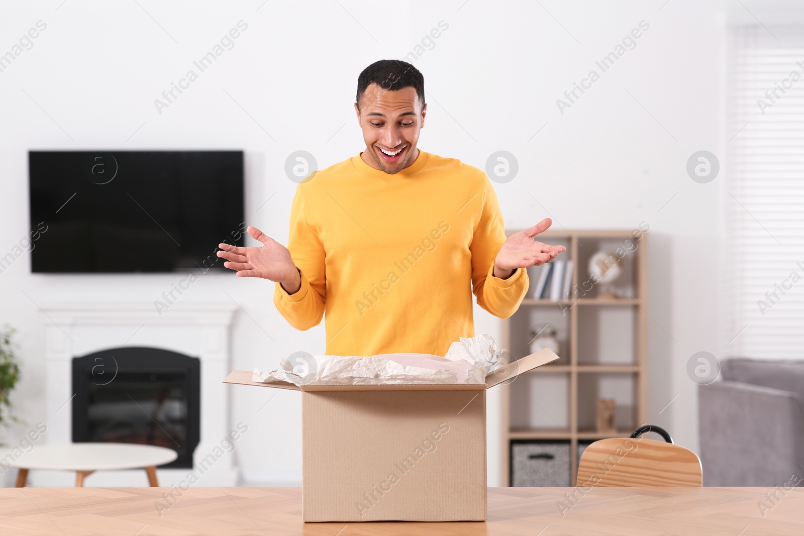 Photo of Emotional young man opening parcel at table indoors. Internet shopping