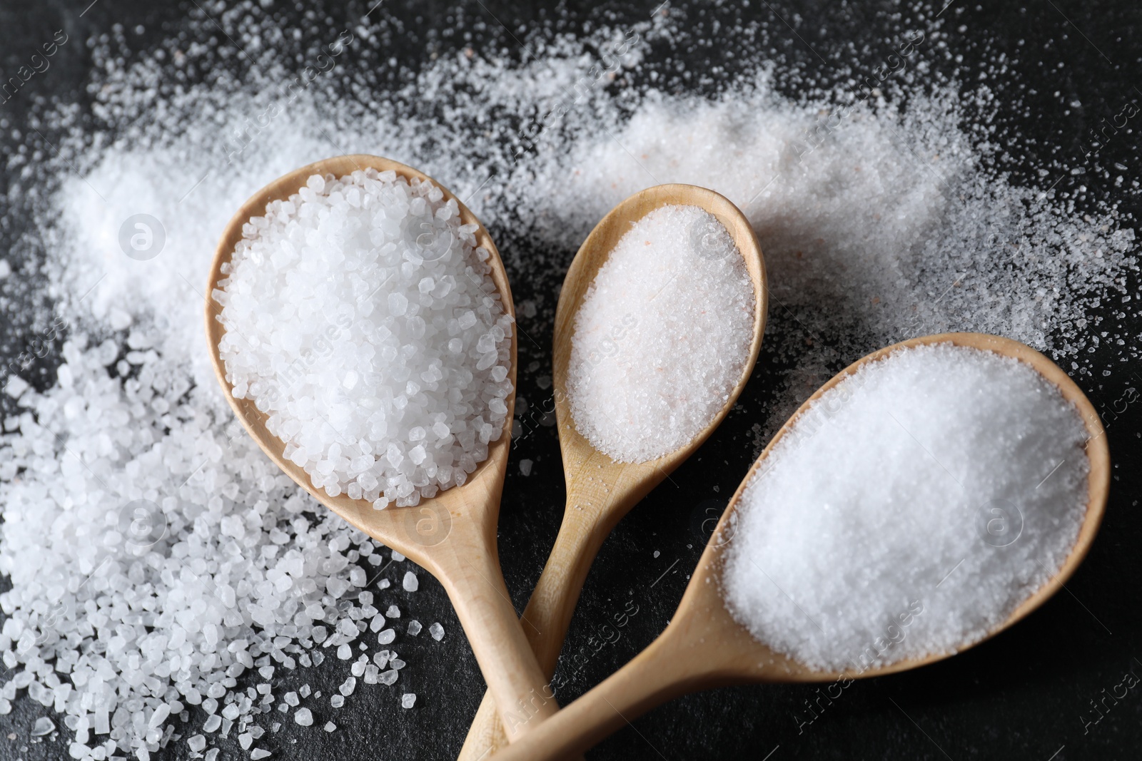 Photo of Organic salt in spoons on black table, flat lay