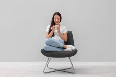Beautiful woman with cup of drink sitting in armchair near light grey wall indoors