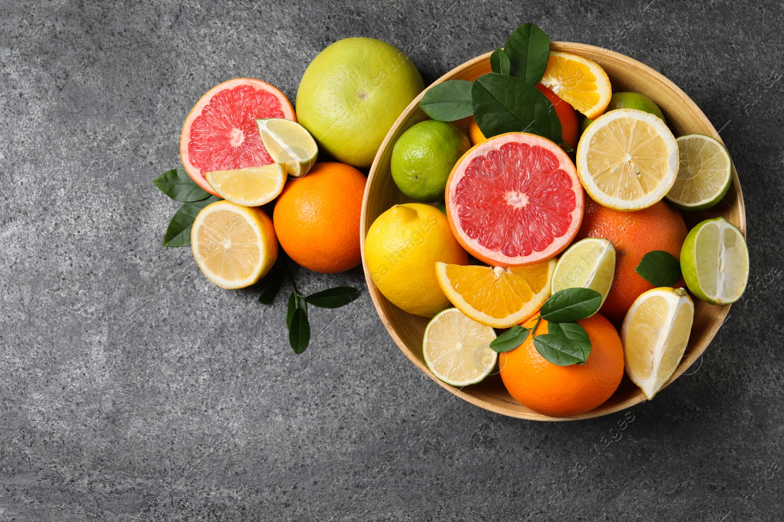 Photo of Different fresh citrus fruits and leaves in bowl on grey textured table, flat lay. Space for text