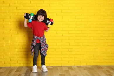 Photo of Cute little boy with skateboard near yellow brick wall indoors, space for text