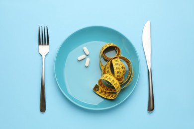 Photo of Plate with weight loss pills, measuring tape and cutlery on light blue background, flat lay