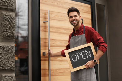 Young male business owner holding OPEN sign near his cafe. Space for text