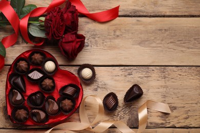 Photo of Heart shaped box with delicious chocolate candies, roses and ribbon on wooden table, flat lay. Space for text