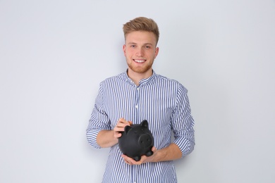 Young man putting coin into piggy bank on light background