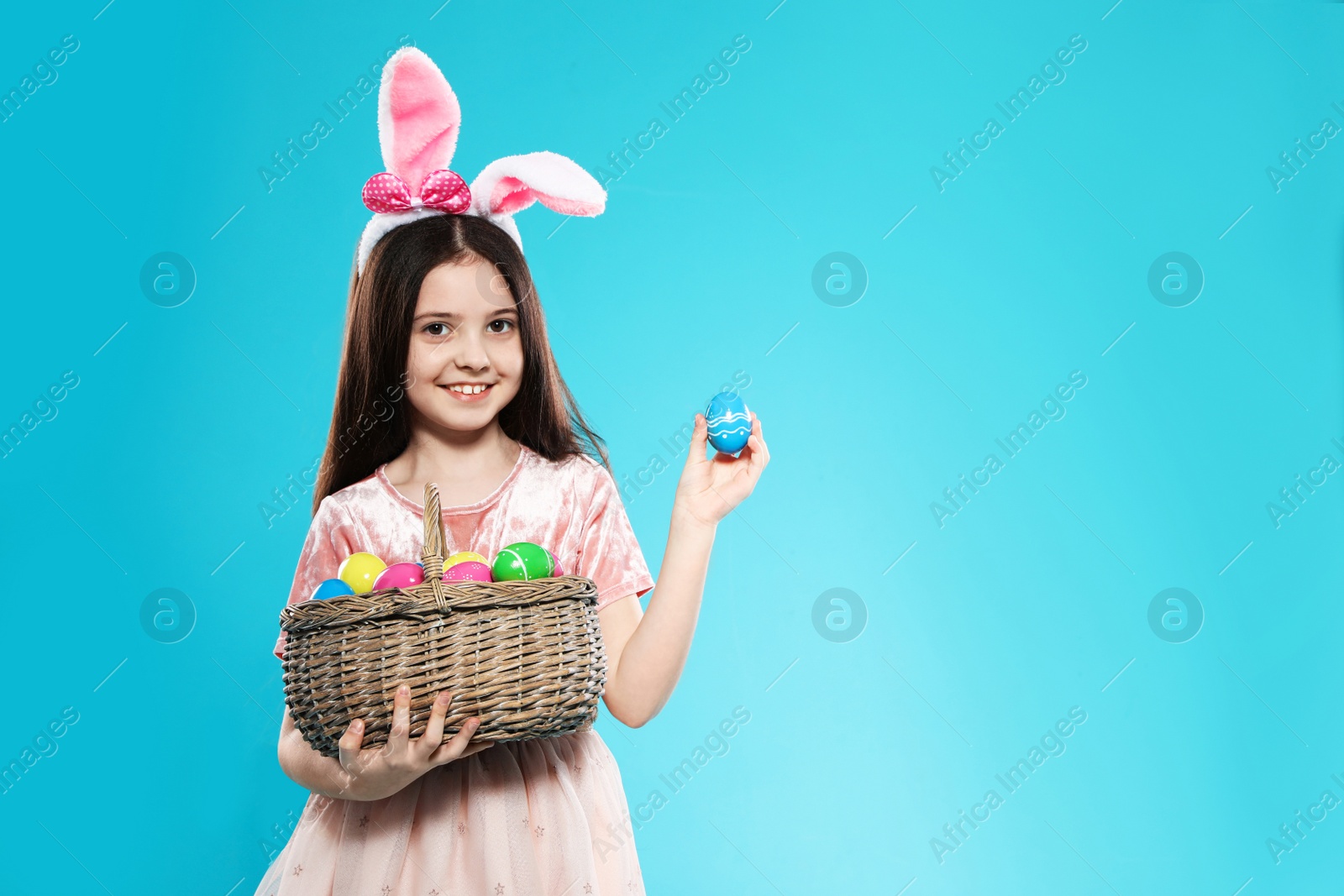 Photo of Little girl in bunny ears headband holding basket with Easter eggs on color background, space for text