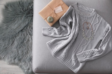 Cozy knitted sweater with jewelry and gift on pouf in living room, flat lay. Space for text