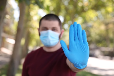 Man in protective face mask showing stop gesture outdoors, focus on hand. Prevent spreading of coronavirus