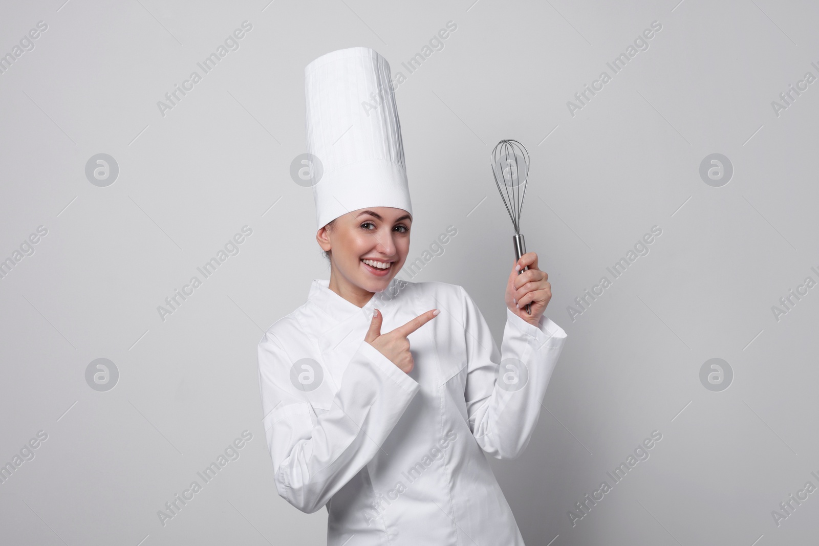 Photo of Happy professional confectioner in uniform pointing at whisk on light grey background