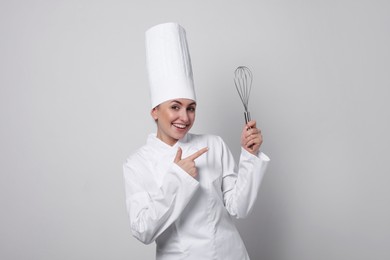 Happy professional confectioner in uniform pointing at whisk on light grey background