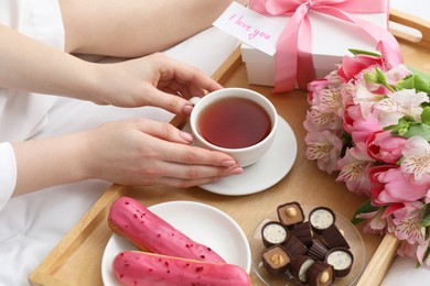 Photo of Tasty breakfast served in bed. Woman with tea, desserts, gift box, flowers and I Love You card at home, closeup