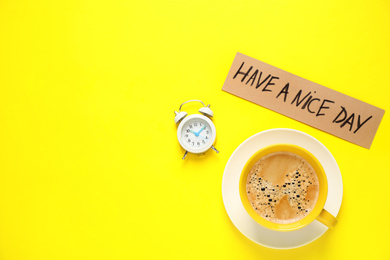 Photo of Delicious morning coffee, alarm clock and card with HAVE A NICE DAY wish on yellow background, flat lay. Space for text