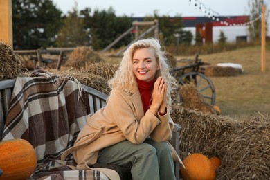 Beautiful woman sitting on wooden bench near pumpkins and hay bales outdoors. Autumn season