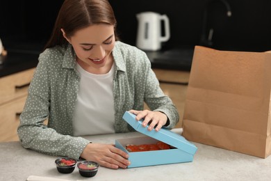 Photo of Beautiful young woman unpacking her order from sushi restaurant at table in kitchen