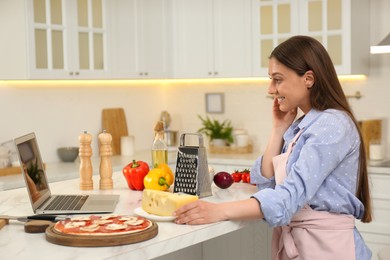 Happy woman making pizza while watching online cooking course via laptop in kitchen