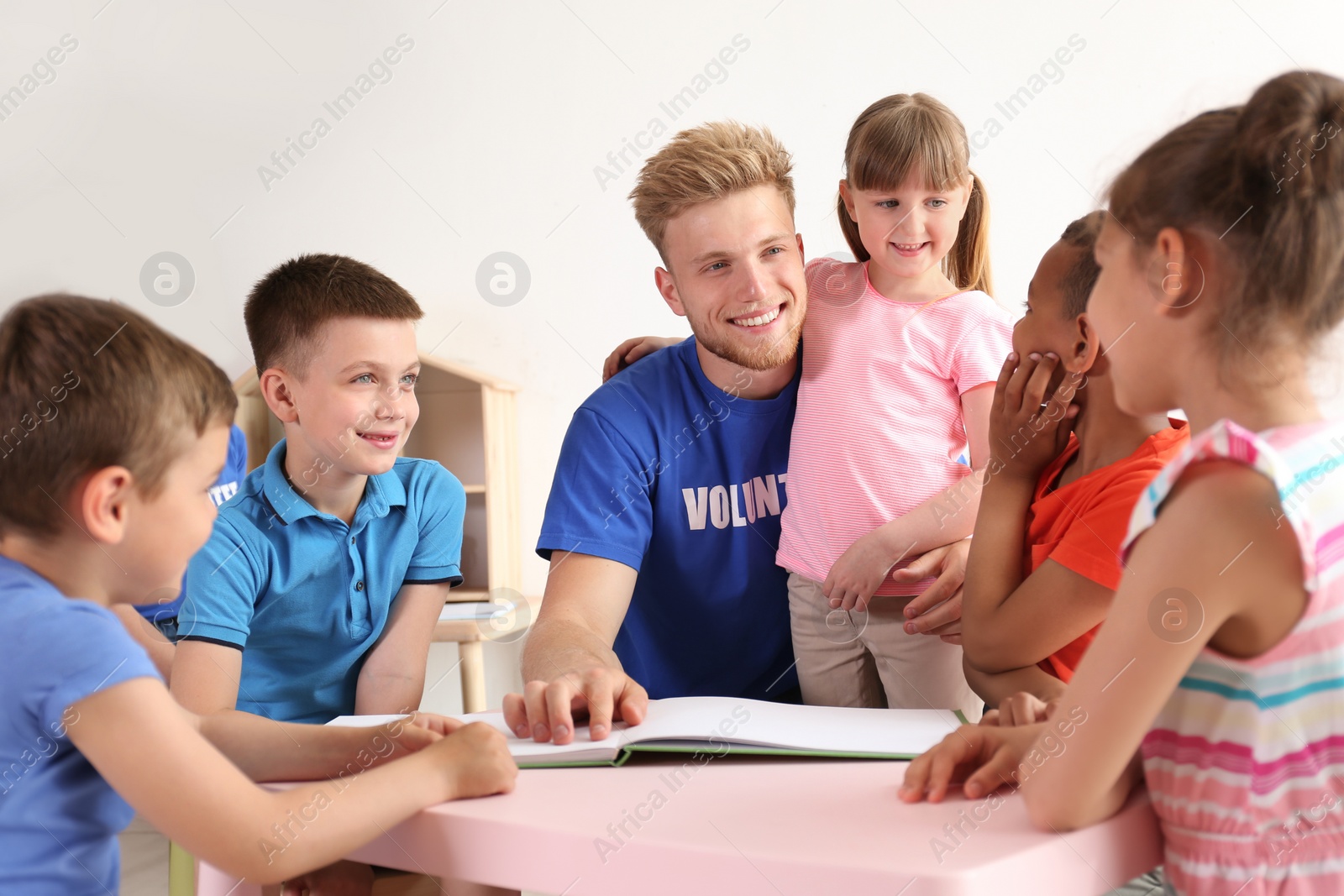 Photo of Young volunteer reading book with children at table indoors