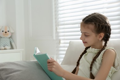 Cute little girl reading book on sofa at home