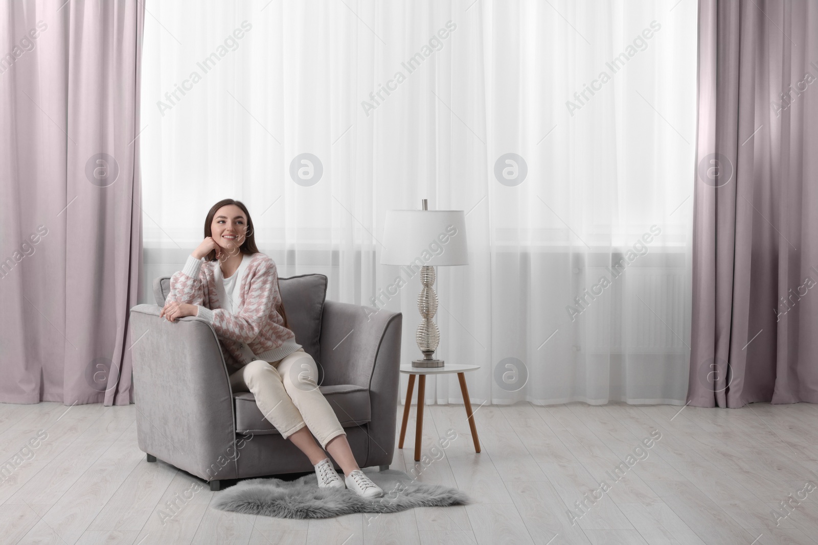 Photo of Woman sitting on armchair near lamp and window with stylish curtains at home. Space for text