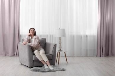 Woman sitting on armchair near lamp and window with stylish curtains at home. Space for text