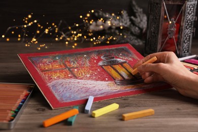 Photo of Woman drawing beautiful lantern with soft pastel at wooden table against blurred lights, closeup